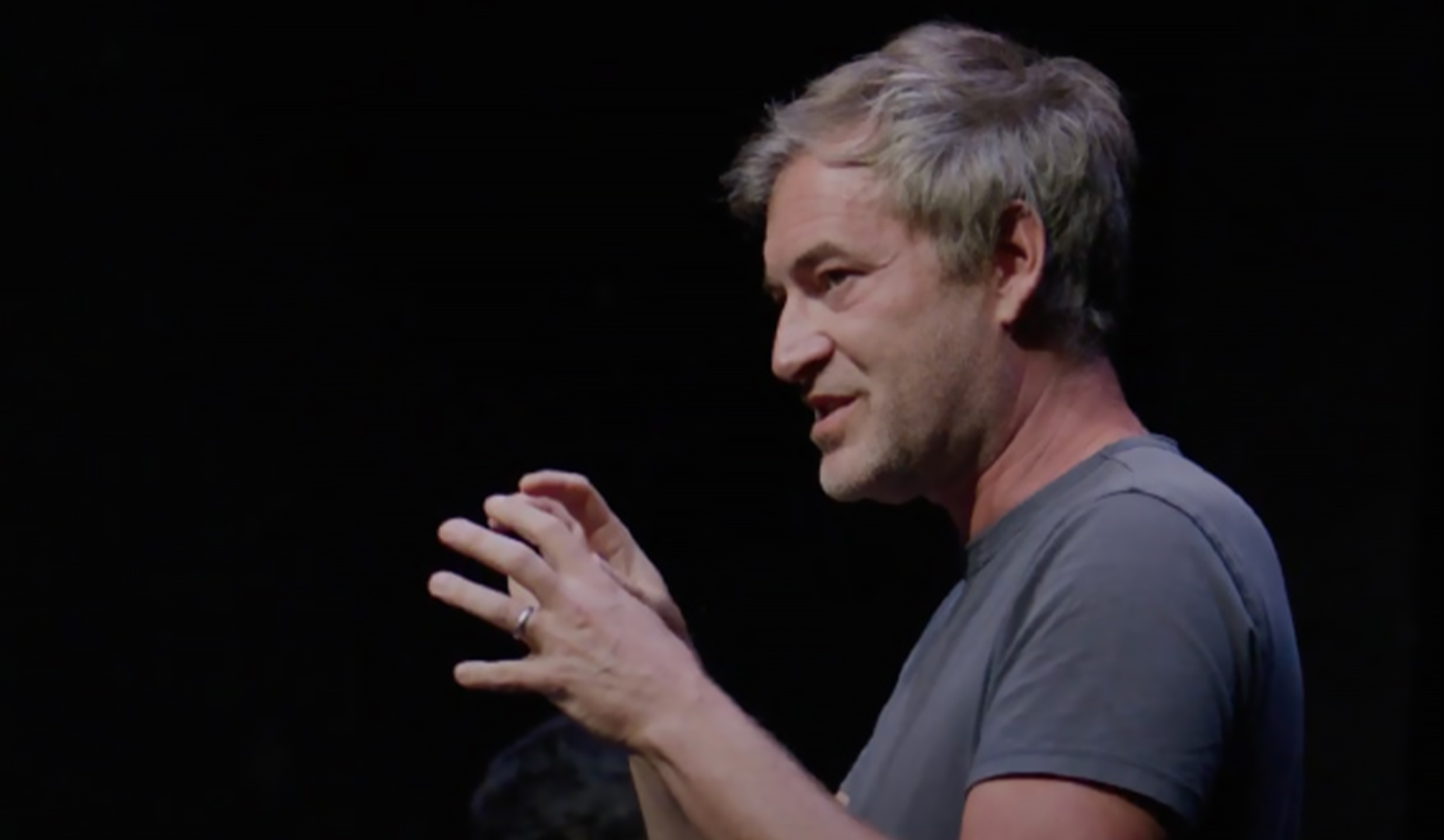 Mark Duplass speaks and gestures with his hands on stage, set against a dark theater setting