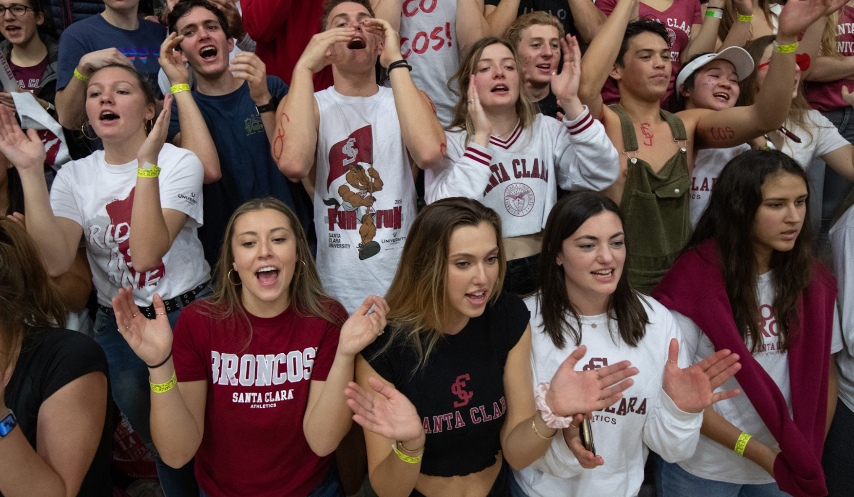 Energetic Santa Clara University students clad in school colors chant and clap in the stands at a sporting event, displaying school spirit and camaraderie.