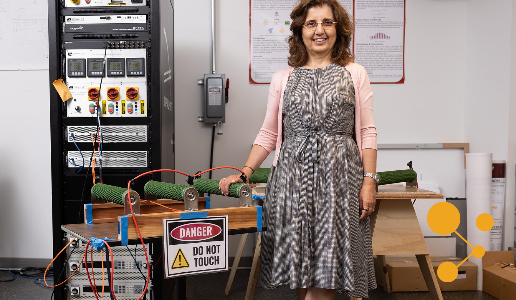 Santa Clara associate professor Maryam Khanbaghi stands confidently in a lab, hand on a microgrid simulator with a 'Danger: Do Not Touch' warning. The background is filled with control panels and safety diagrams, highlighting a sophisticated electrical engineering research setting.