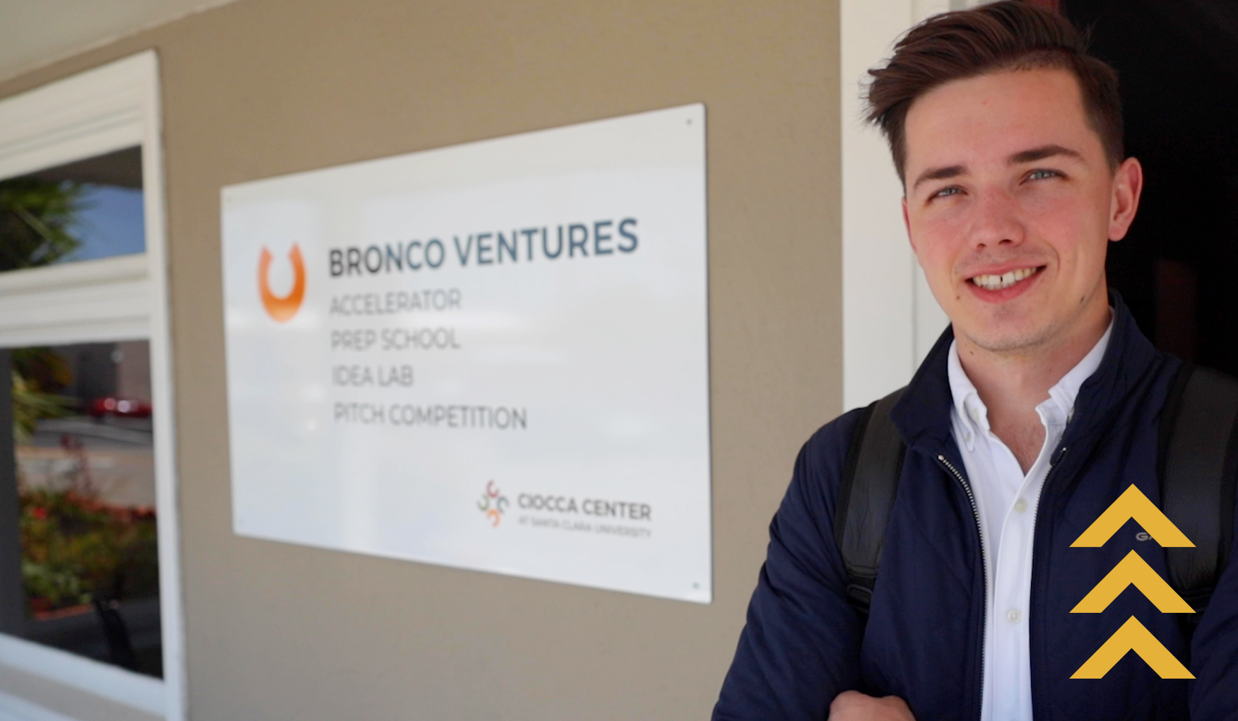 A Santa Clara student stands in front of the 'Bronco Ventures' sign. He is dressed in business casual attire with a backpack, with his arms crossed. The sign indicates an accelerator, prep school, idea lab, and pitch competition at the Ducca Center.